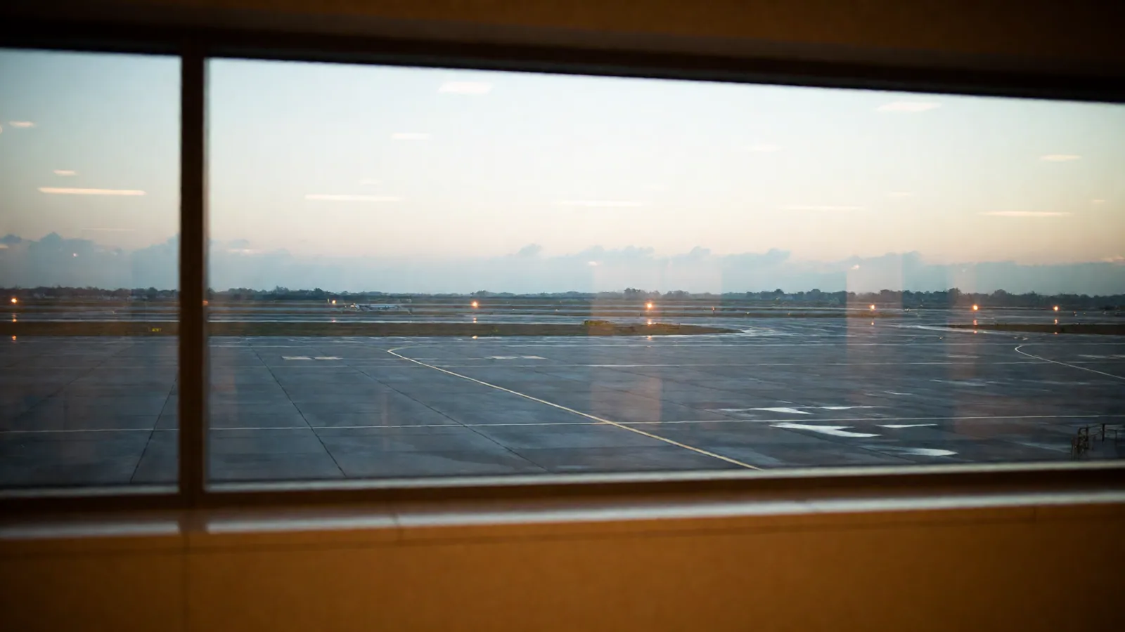 A long terminal window looking out over an empty airfield at dusk, with the runway lights just come on